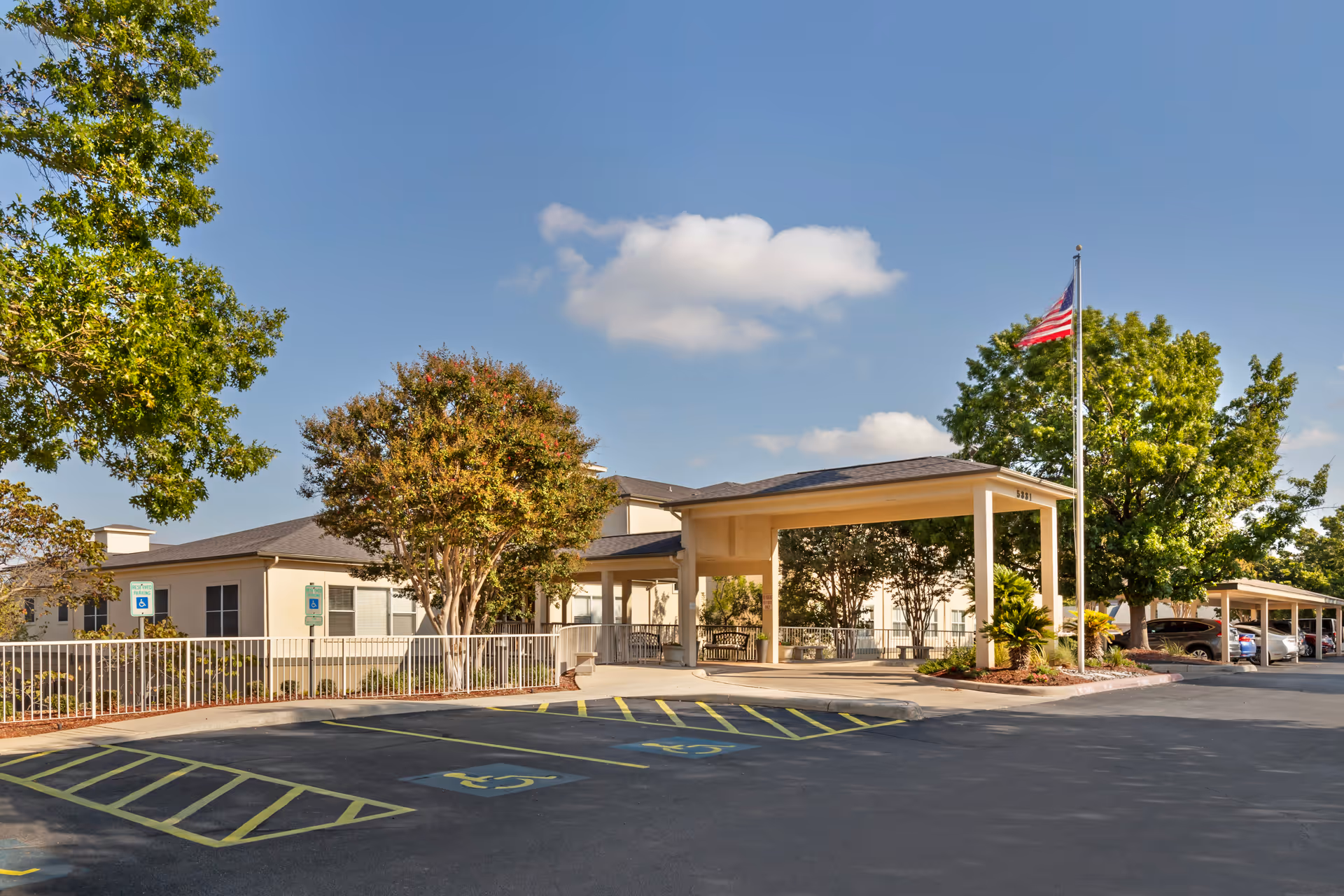 Front entrance of a senior living facility with a covered drive-through, American flag, accessible parking spaces, and surrounding trees.