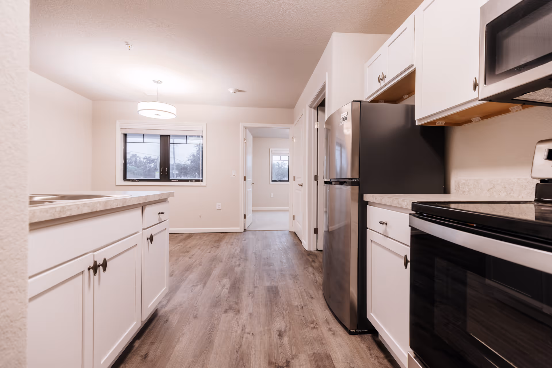 Interior view of a modern kitchen with white cabinets, a stainless steel refrigerator, stove, and microwave. The kitchen has light-colored countertops and wood flooring. A window and a hanging light fixture are visible in the adjacent dining area, with doorways leading to other rooms.