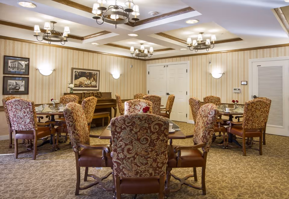 A dining room with several round tables surrounded by upholstered chairs with floral patterns. The room features beige patterned carpet, striped wallpaper, and multiple light fixtures including chandeliers and wall sconces. There is a piano against the far wall and framed pictures hanging above it.