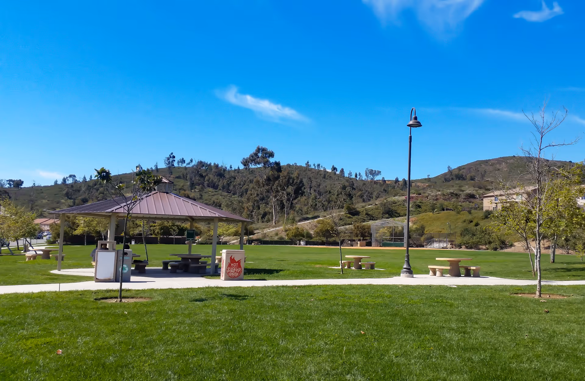 A sunny outdoor park area with green grass, a covered picnic pavilion with benches and tables, a lamppost, and several small trees. Hills and a clear blue sky are visible in the background.