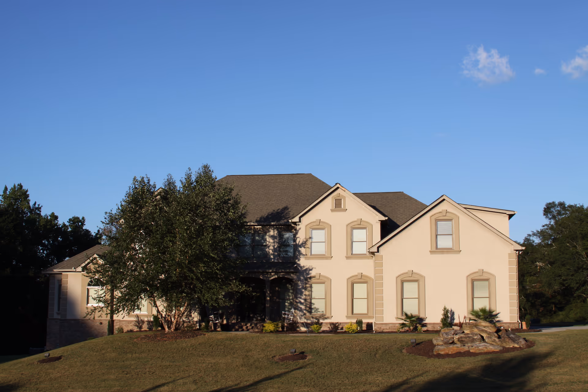 A large two-story beige house with multiple windows and a dark roof, situated on a grassy hill with trees around it under a clear blue sky.