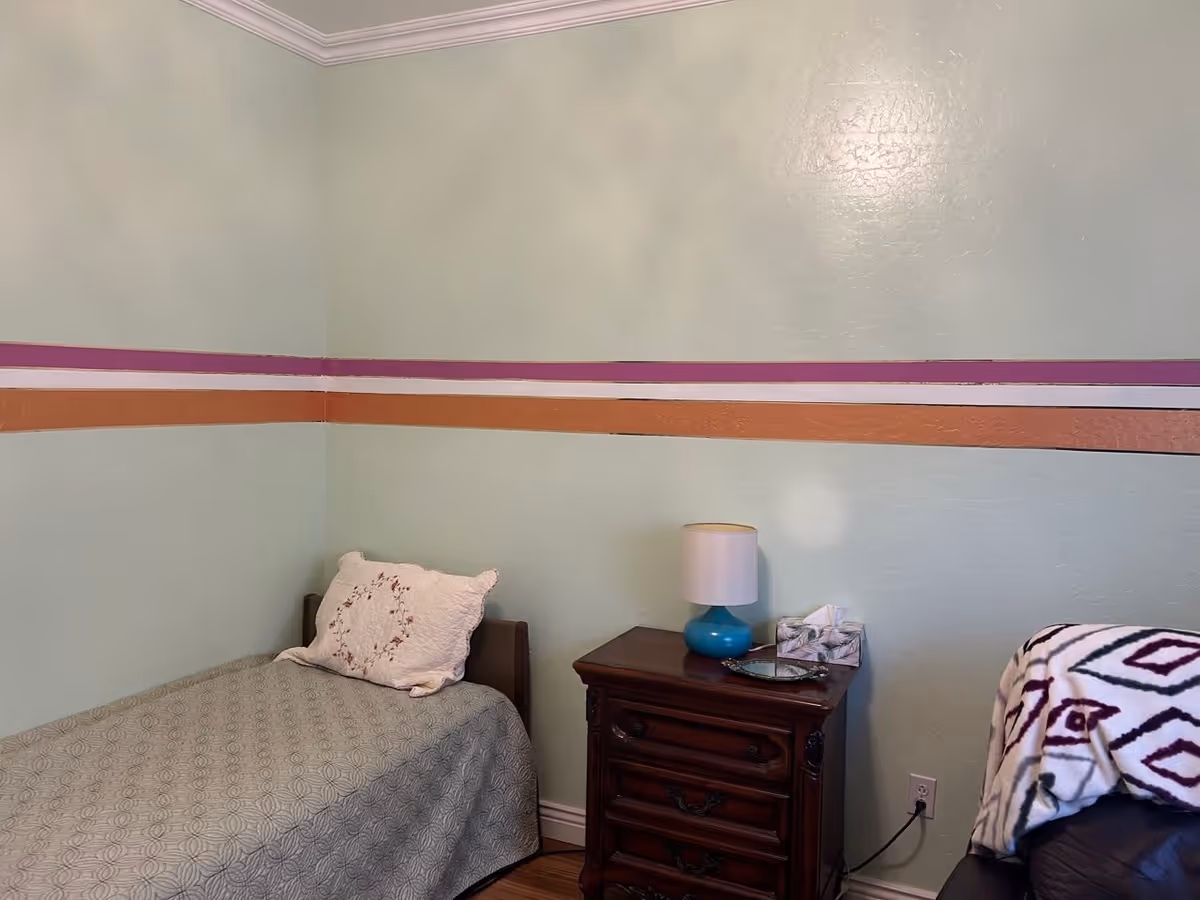 A small bedroom with a single bed covered in a gray patterned bedspread and a decorative pillow. Next to the bed is a dark wooden nightstand with a blue lamp, a tissue box, and a small tray. The walls are painted light green with horizontal stripes in purple, white, and orange. A blanket with a geometric pattern is draped over a piece of furniture on the right side.