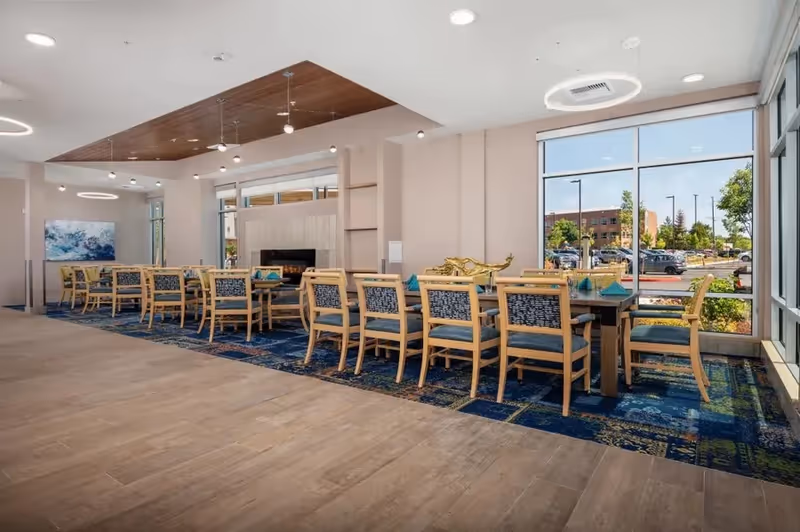 A spacious dining room with a long table surrounded by wooden chairs with blue patterned cushions. The room features large windows letting in natural light, a modern fireplace, a blue patterned carpet under the table, and contemporary ceiling lights. Outside the windows, a parking lot and greenery are visible.