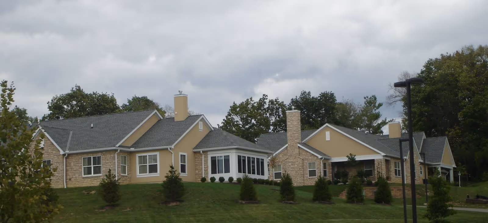 Exterior view of a single-story senior living facility building with beige walls, stone accents, multiple windows, and a gray shingled roof, surrounded by green grass and small trees under a cloudy sky.
