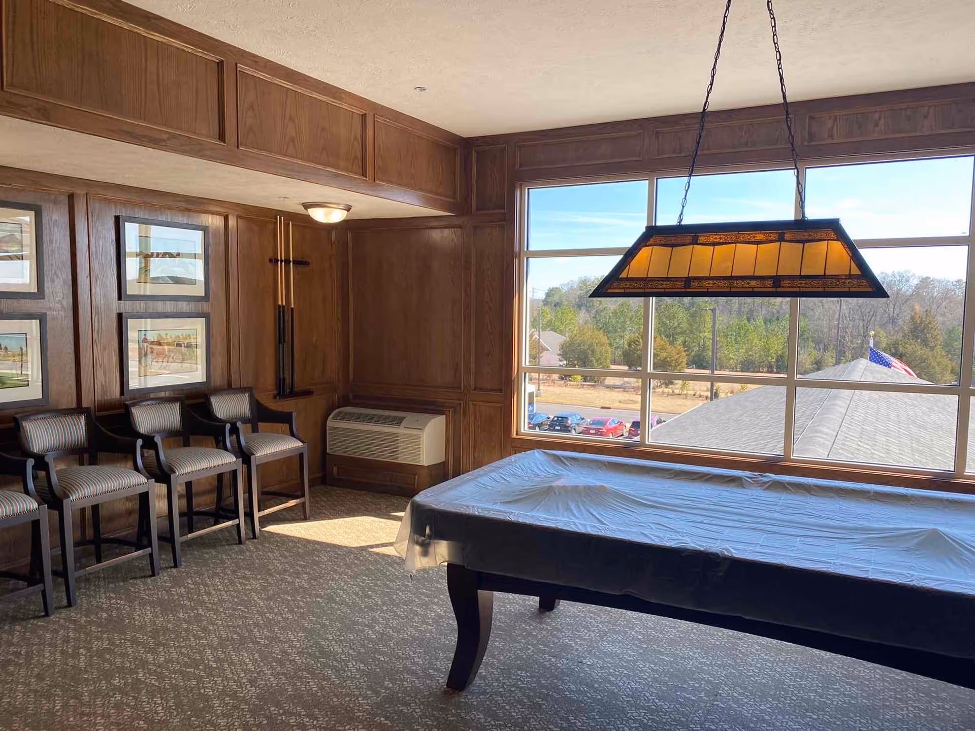 A room with wood-paneled walls featuring framed artwork, three striped upholstered chairs lined up against the wall, a pool table covered with a protective cloth, a hanging rectangular light fixture above the pool table, a large window showing an outdoor view with trees, parked cars, and an American flag.