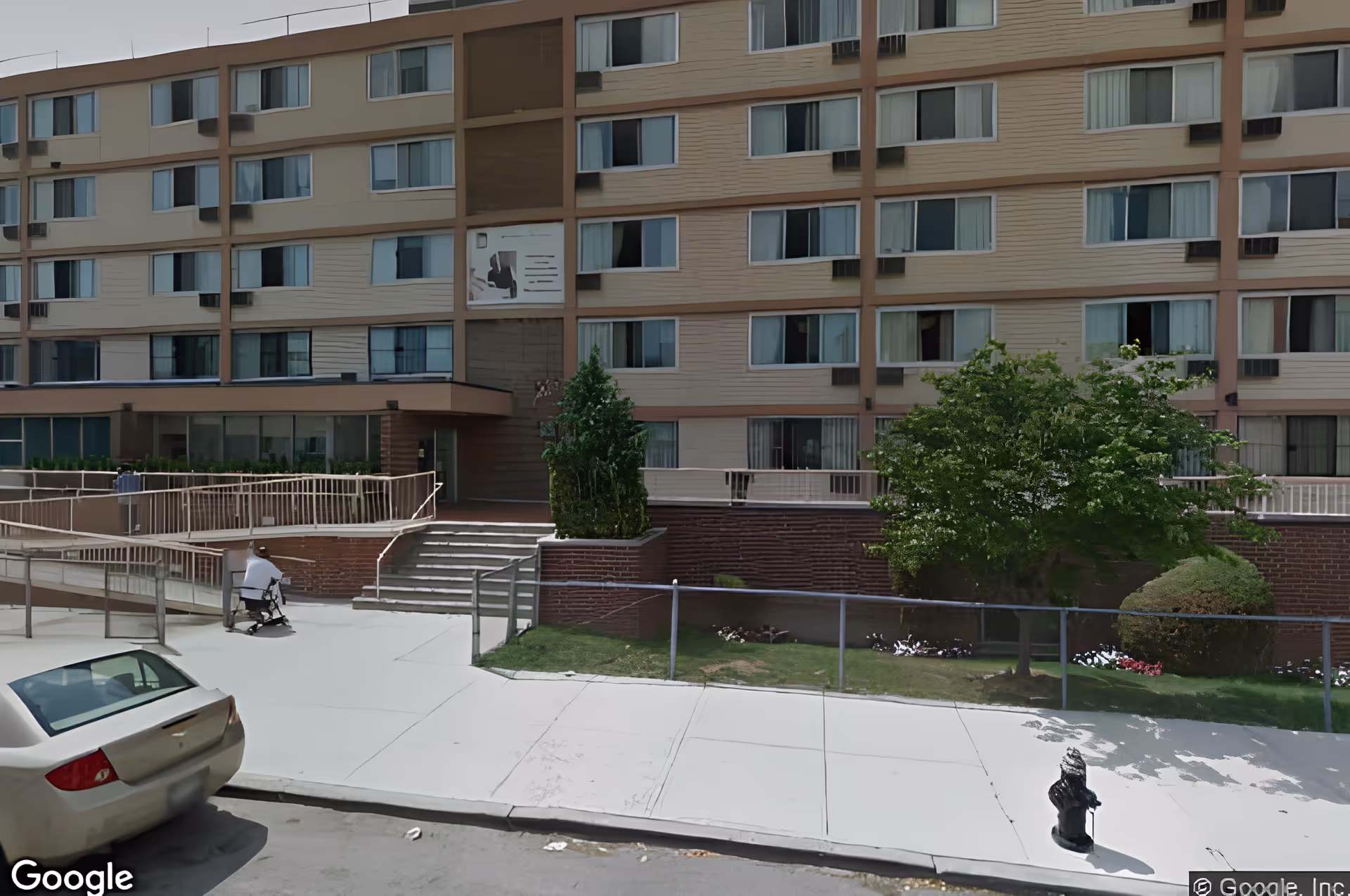 Exterior view of Amber Court of Brooklyn, a multi-story senior living facility with numerous windows. There is a sidewalk and a ramp leading to the entrance, some greenery including bushes and a tree, a parked car, and a person using a mobility scooter near the entrance.
