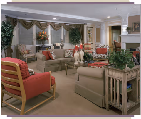 A cozy living room with beige sofas and red cushioned chairs arranged around a glass coffee table. The room features large windows with green drapes, decorative plants, framed artwork on the walls, and a fireplace on the right side. The space is warmly lit with ceiling lights and table lamps.