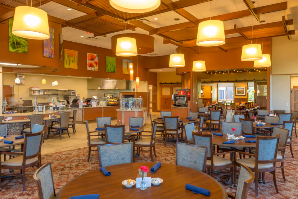 Spacious, well-lit dining room with round tables and chairs, a serving counter and hanging pendant lights.