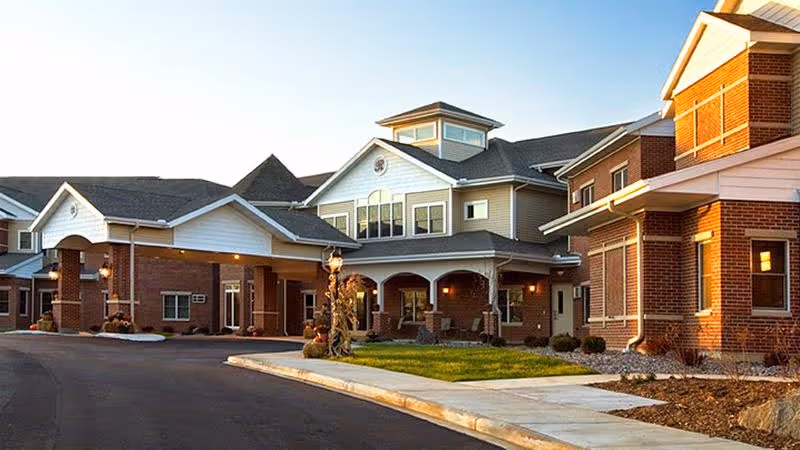 Exterior view of a senior living facility building with brick and siding, featuring a covered entrance with columns, well-maintained landscaping, and a paved driveway.