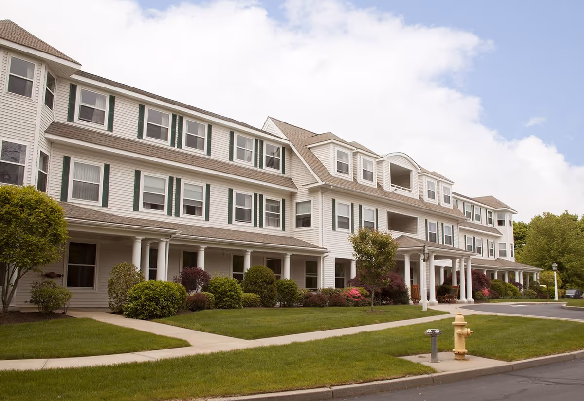 Exterior view of a large, three-story senior living facility building with white siding, multiple windows, and a covered entrance supported by white columns. The building is surrounded by well-maintained green lawns, shrubs, and small trees under a partly cloudy sky.