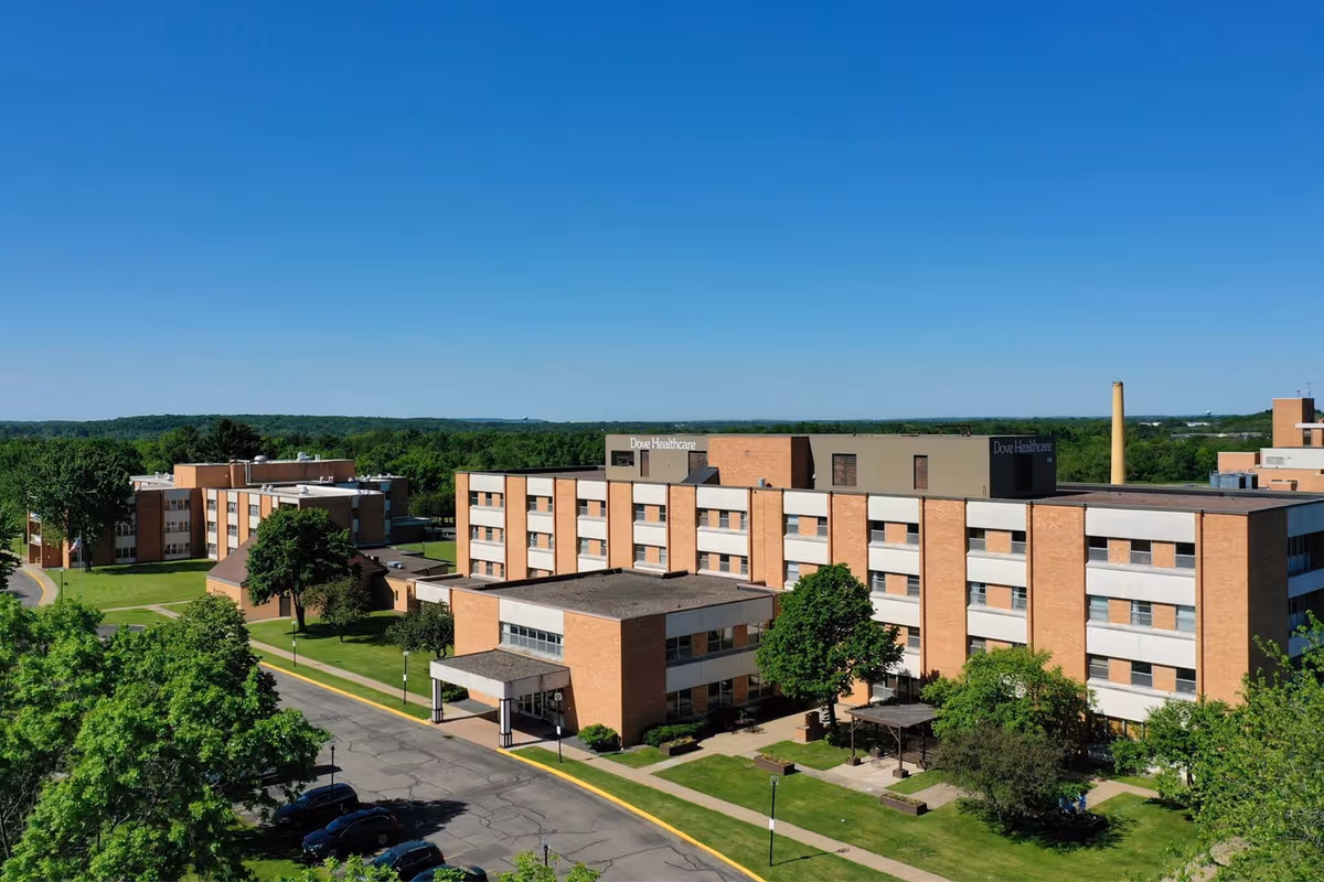Aerial view of a multi-story brick senior healthcare building with lawns, trees, and a driveway under a clear blue sky.