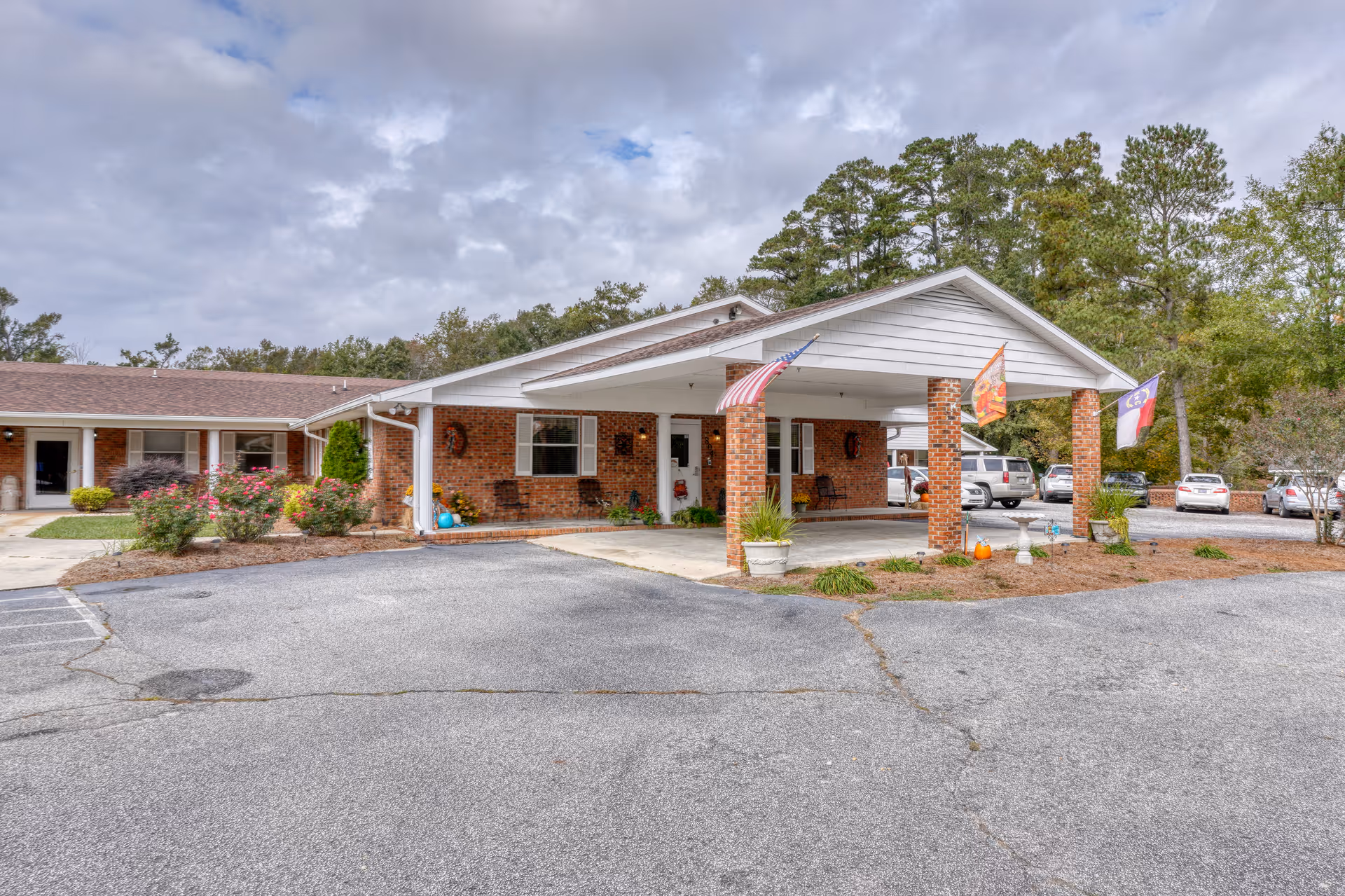 Exterior view of Bladen East Health and Rehab facility showing a single-story brick building with a covered entrance supported by brick columns. There are three flags displayed near the entrance, several parked cars, landscaped bushes, and trees in the background under a cloudy sky.
