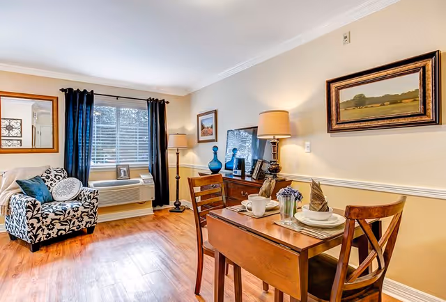 A cozy living and dining area with wooden flooring, a small wooden dining table set for two with plates, cups, and napkins, a patterned armchair with cushions near a window with dark curtains, a wooden sideboard with a lamp, decorative items, and framed pictures on the wall.