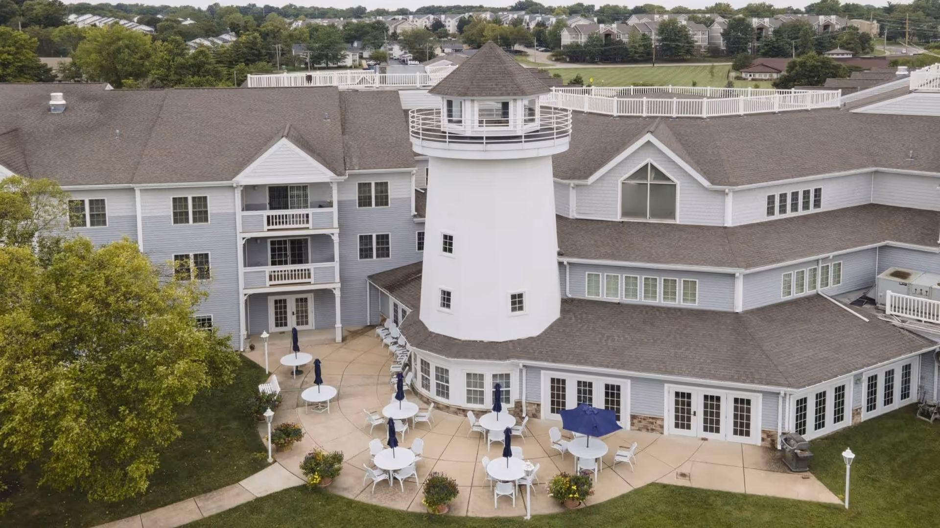 Aerial view of Cape Albeon Independent Living facility showing a large building with a distinctive white lighthouse structure in the center. The building has multiple windows and balconies. Outside, there is a patio area with several round tables, chairs, and closed umbrellas. Surrounding the building are green lawns, trees, and a residential neighborhood in the background.