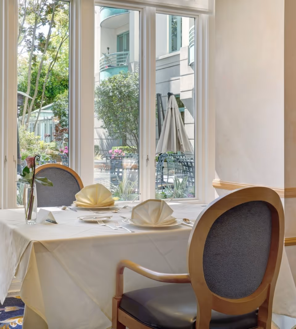 A neatly set dining table with folded napkins and chairs beside large windows overlooking a courtyard.