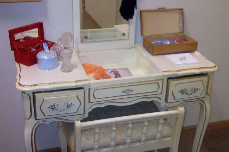 A vintage-style vanity table with a mirror, featuring a red jewelry box, glass containers, and a wooden box with items inside. A cushioned chair is placed in front of the vanity.