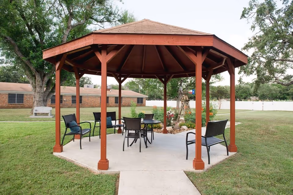 A wooden gazebo with a hexagonal roof situated on a concrete pad in a grassy outdoor area. Inside the gazebo are several black metal chairs with cushions and a round table. In the background, there are trees, a brick building, a white fence, and a small garden with flowers and a decorative windmill.