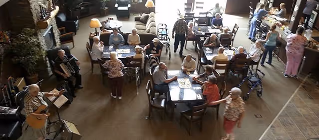 A large common area in a senior living facility with multiple tables where elderly residents are seated and engaged in activities such as playing board games and socializing. Some residents are using walkers and wheelchairs. There is a musician playing an accordion and another playing a guitar. The room is well-lit with natural light coming from large windows, and there are sofas and lamps in the background.
