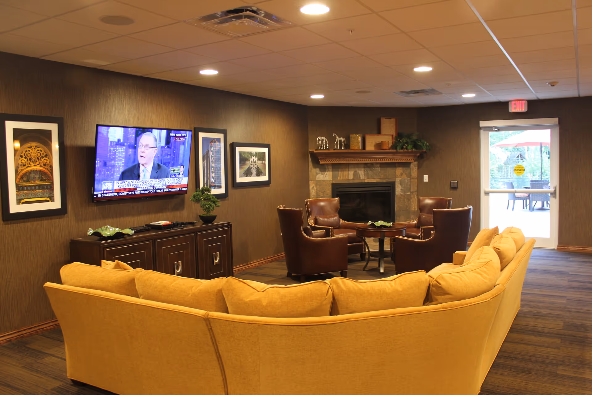 A cozy living room area in Cahaba Ridge Retirement Community featuring a large yellow sectional sofa facing a wall-mounted flat-screen TV. The TV is flanked by framed artwork. There is a stone fireplace with a wooden mantle decorated with small sculptures and plants. Four brown leather chairs surround a small round table near the fireplace. A glass door leads to an outdoor patio with seating and an umbrella.