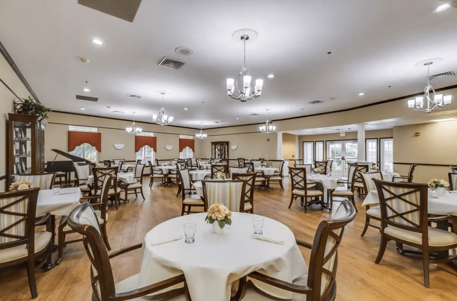A spacious dining room with multiple round tables covered with white tablecloths, each set with glasses and napkins. The room features wooden chairs with cushioned seats, chandeliers hanging from the ceiling, large windows with red valances, and a grand piano in the corner.