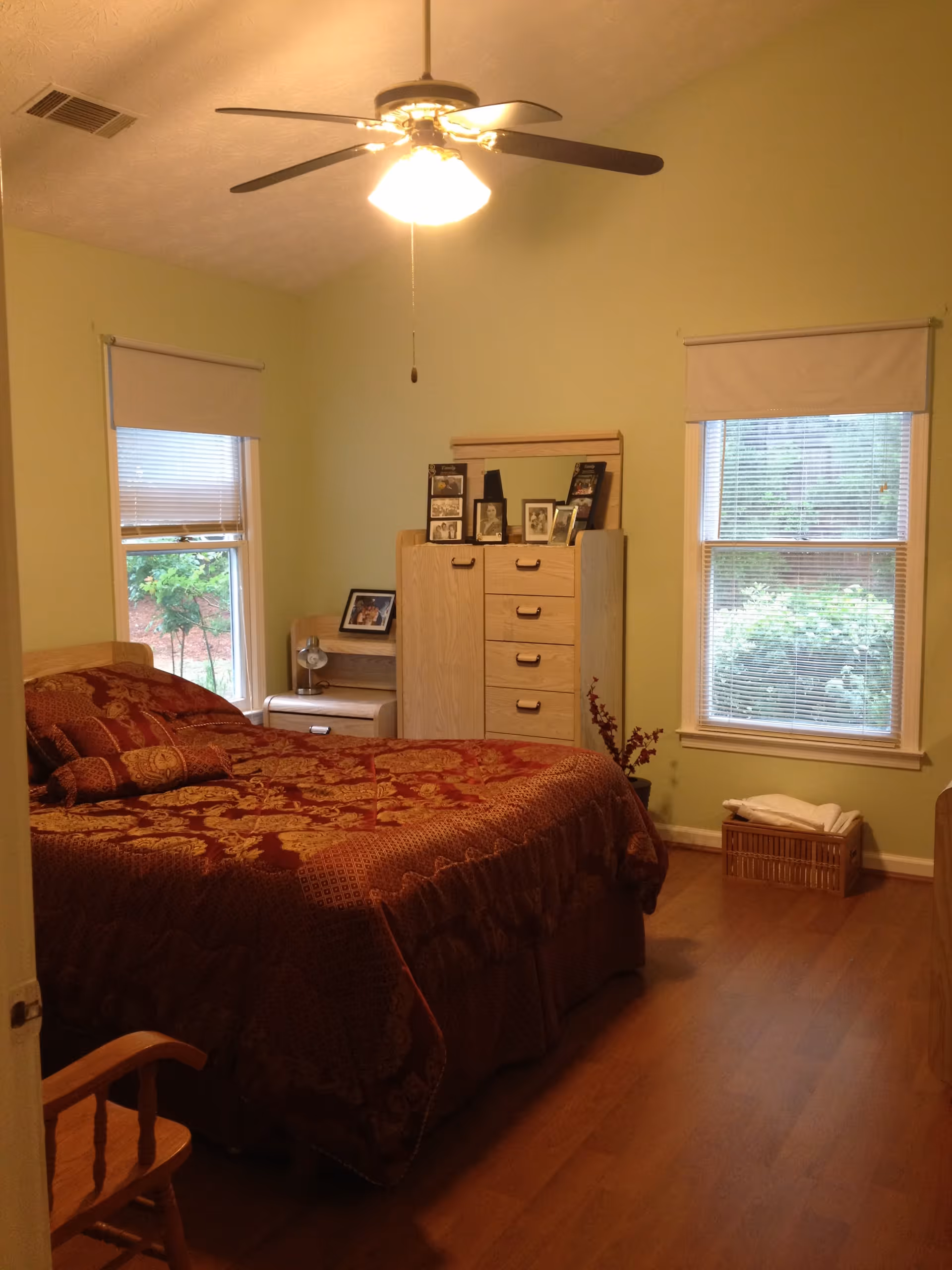 A cozy bedroom with light green walls, a ceiling fan with a light, two windows with blinds, a wooden bed with a red and gold patterned bedspread, a wooden dresser with framed photos on top, a small nightstand with a lamp, and a wooden chair in the corner.