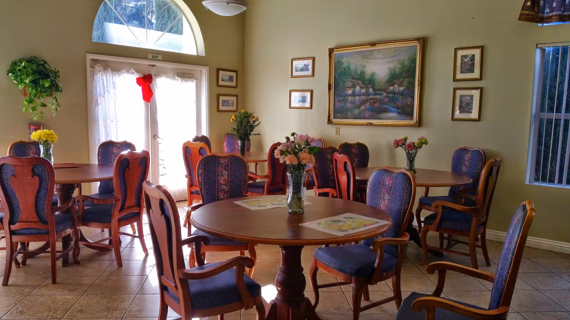 A cozy dining room with several round wooden tables surrounded by upholstered chairs with wooden frames. Each table has a vase with flowers. The room has tiled floors, light-colored walls adorned with framed paintings, and a large arched window with sheer curtains letting in natural light.