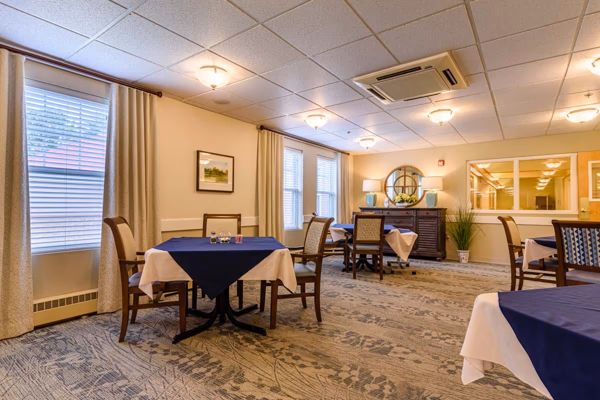 A dining room with several tables covered with white and navy blue tablecloths, surrounded by wooden chairs with cushioned seats. The room has beige walls, large windows with curtains, a patterned carpet, and a sideboard with a round mirror and two lamps on top.