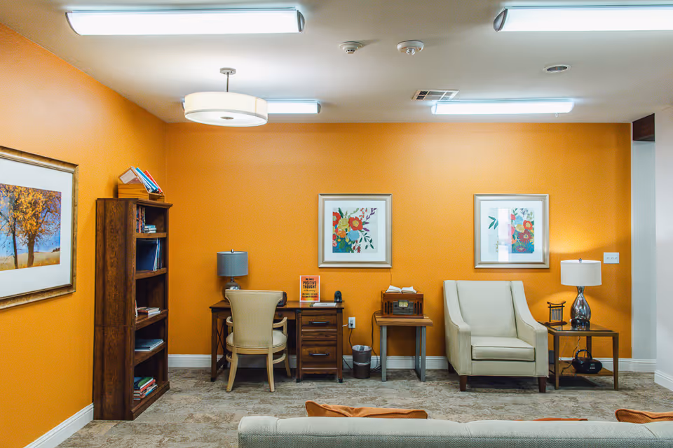 A cozy interior room with orange walls featuring a wooden bookshelf filled with books, a desk with a chair and a lamp, a small table with a vintage radio, and a comfortable armchair next to a side table with a lamp. Two framed floral artworks hang on the wall above the armchair and table.