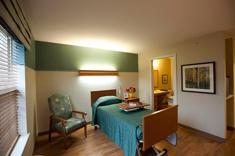 A furnished private bedroom in a senior care facility with a single bed covered in a green quilt, a bedside table, an armchair, a window, and an open door to a bathroom.