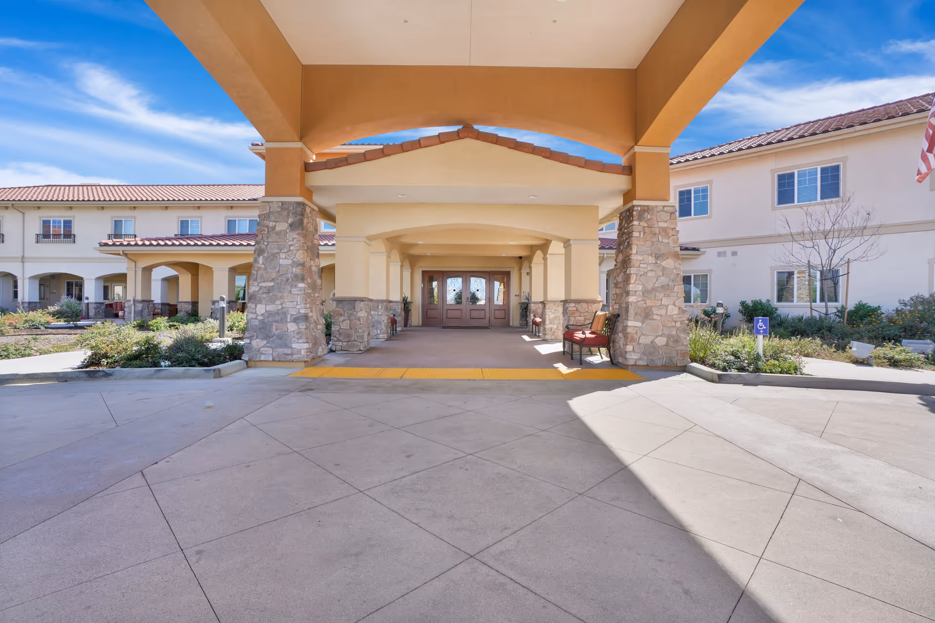 Entrance of a senior living facility with a covered driveway supported by stone pillars, beige walls, and a tiled roof. There are chairs placed near the entrance, landscaped greenery on both sides, and a clear blue sky above.