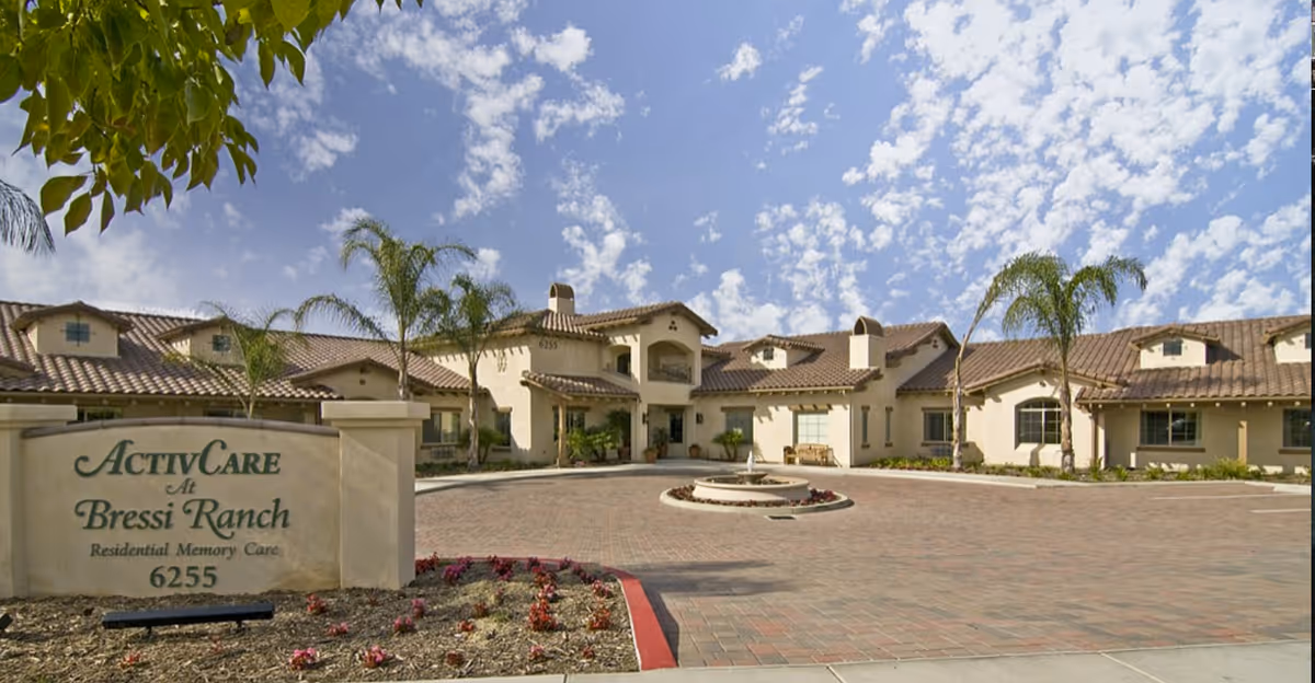 Exterior view of ActivCare at Bressi Ranch, a residential memory care facility, showing a large building with beige walls, a tiled roof, palm trees, and a circular driveway with a small fountain in the center under a partly cloudy sky.