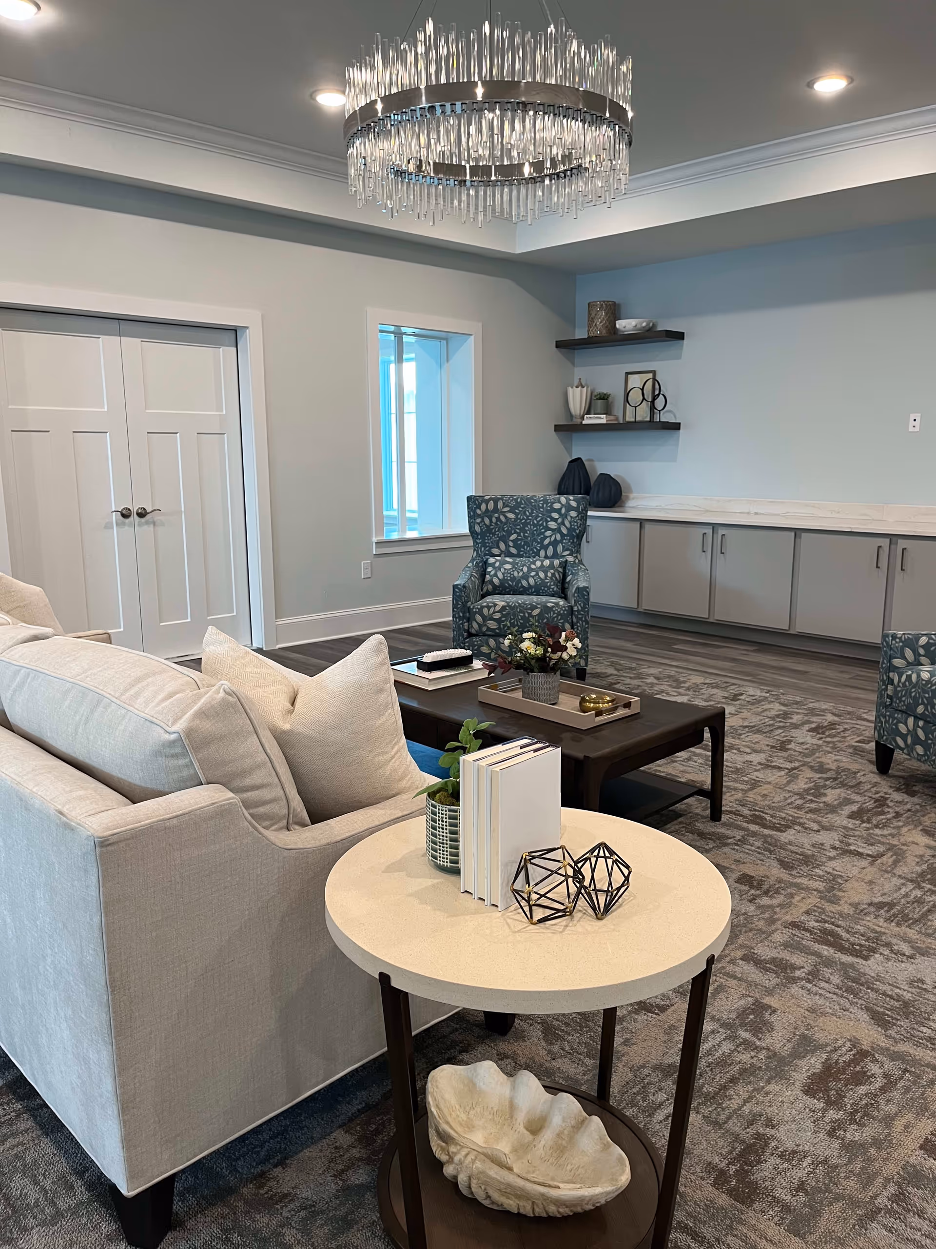 A modern living room area with a light beige sofa, a round side table with decorative items including books and geometric sculptures, two patterned armchairs, a dark wooden coffee table with a flower arrangement, built-in cabinets along the wall, floating shelves with decorative items, a window, and a large crystal chandelier hanging from the ceiling.