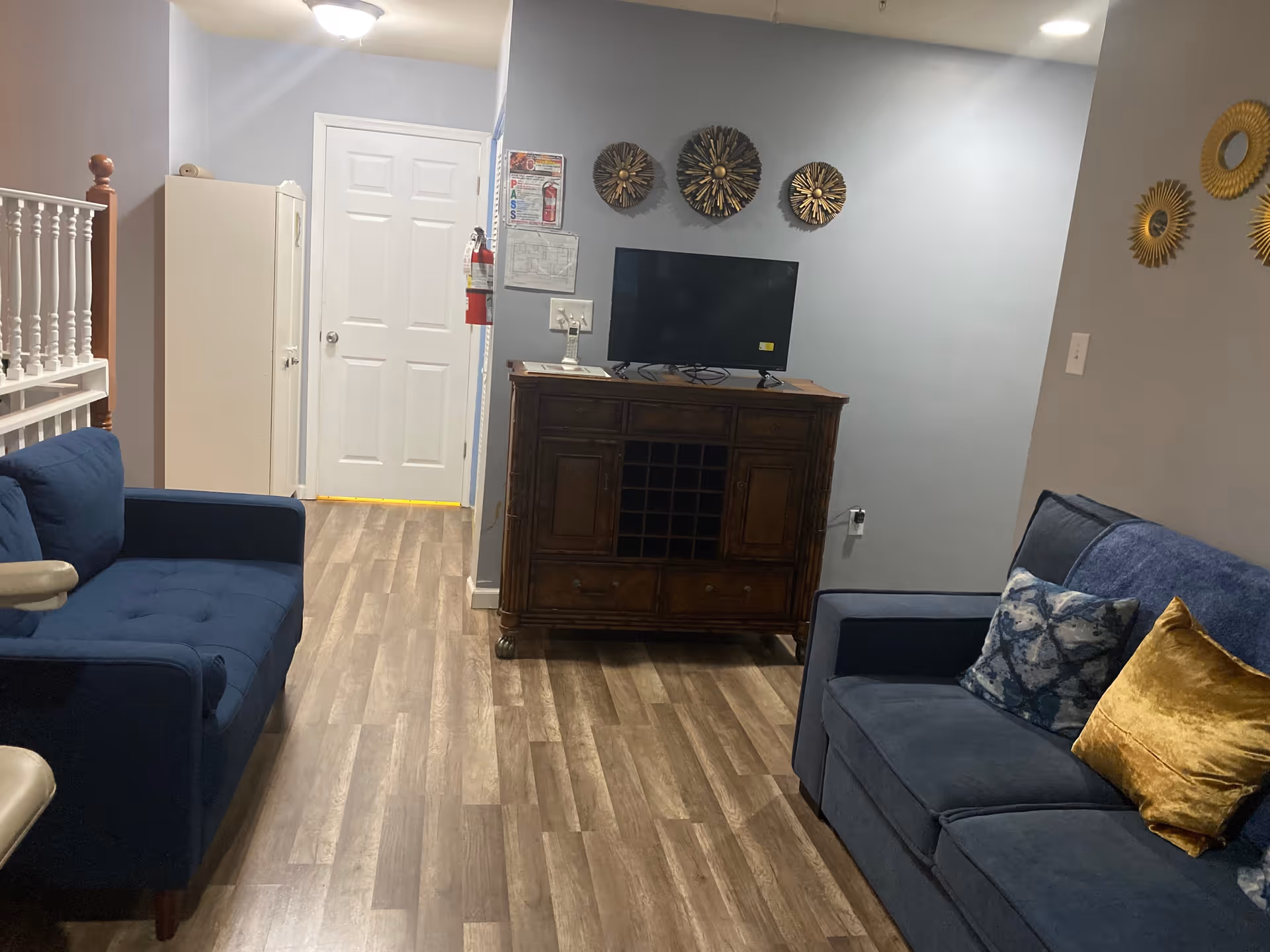 Interior view of a living room area with two blue upholstered sofas, one on the left and one on the right with decorative pillows. A wooden cabinet with a flat-screen TV on top is against the far wall, which is painted light blue. Above the TV are three decorative wall hangings. The floor is wood laminate, and there is a white door and a white cabinet in the background. A fire extinguisher is mounted on the wall near the door.