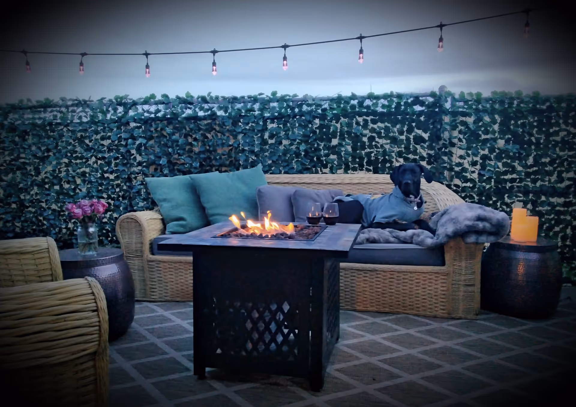 Outdoor patio area with wicker furniture including a sofa and chair, a fire pit table with flames, two glasses of red wine, a small table with a vase of pink flowers, string lights hanging above, and a black dog wearing a gray sweater lying on the sofa with cushions and a blanket.