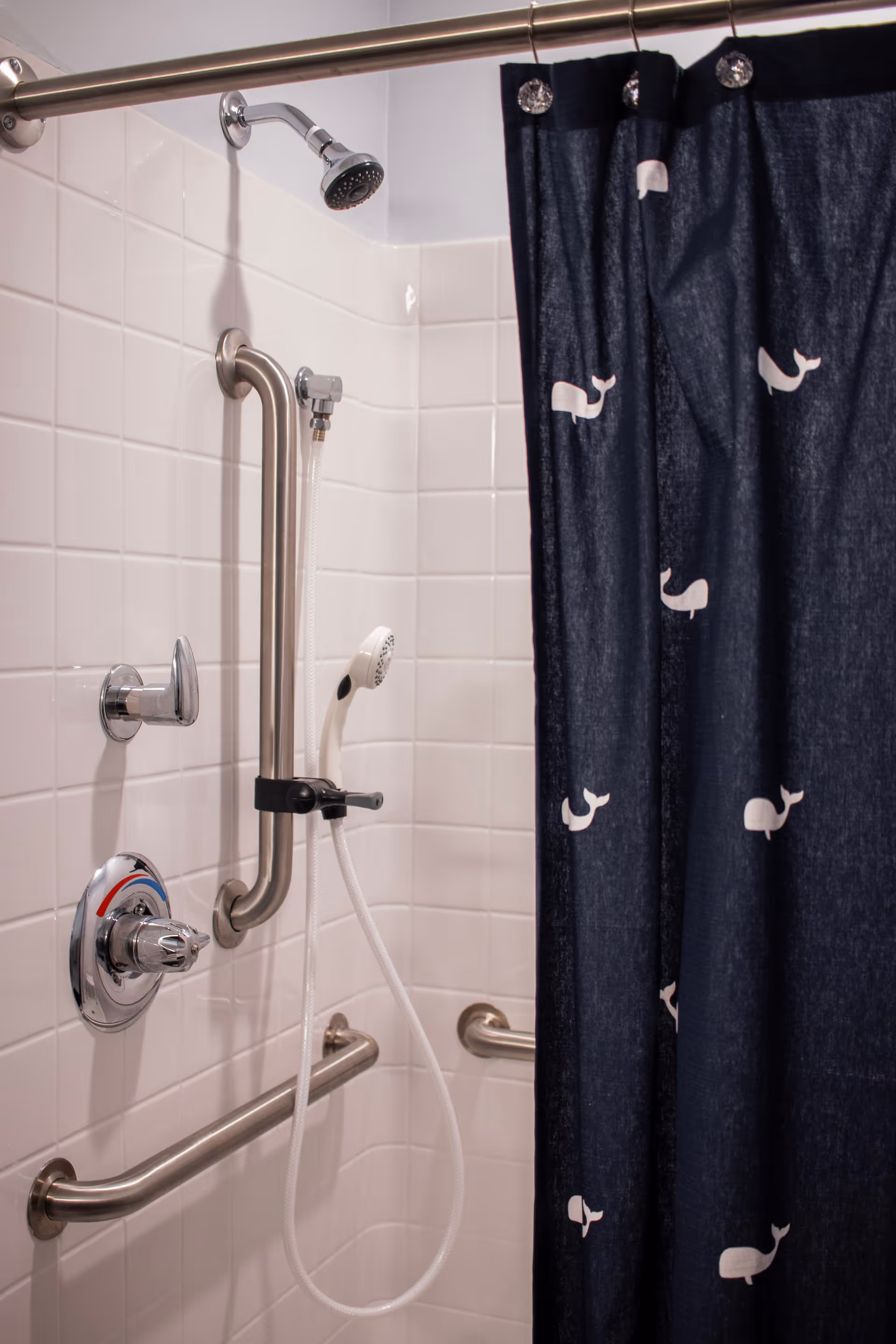 A shower area with white tiled walls, equipped with a fixed showerhead and a handheld showerhead attached to a vertical grab bar. There are additional horizontal grab bars for support. A dark blue shower curtain with white whale patterns is partially drawn to the side.