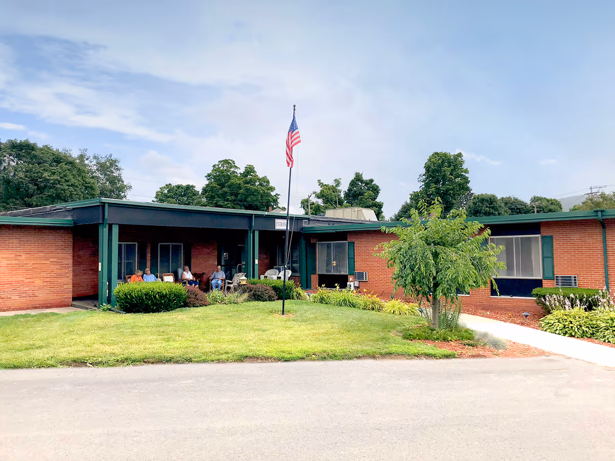 Exterior view of a single-story brick building with a green roof and trim, featuring a small covered porch area where several people are seated. There is a well-maintained lawn with bushes and a tree in front, and an American flag on a flagpole near the center of the lawn. The sky is partly cloudy.