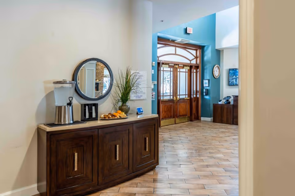Interior view of a senior living facility entrance area with wooden double doors, a wooden cabinet with a water dispenser, a plate of pastries, a vase with green plants, and a round mirror on the wall. The floor is tiled and the walls are painted in light and teal colors.