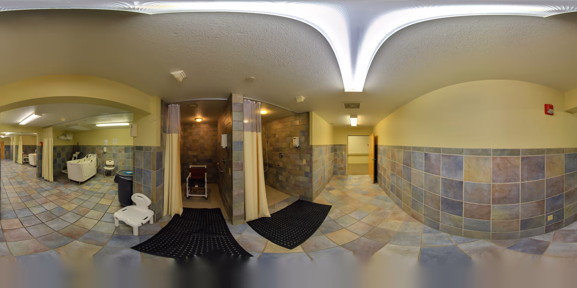 A tiled room with two accessible shower stalls separated by curtains, each equipped with a shower chair. The room has tiled floors and walls with a mix of beige, gray, and brown tiles. There is a white plastic chair and a large white sink with a chair nearby. The lighting is warm and the ceiling is white with a curved design.