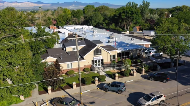 Aerial view of Draper Rehabilitation and Care Center building surrounded by trees and parked cars, with mountains visible in the background under a clear sky.