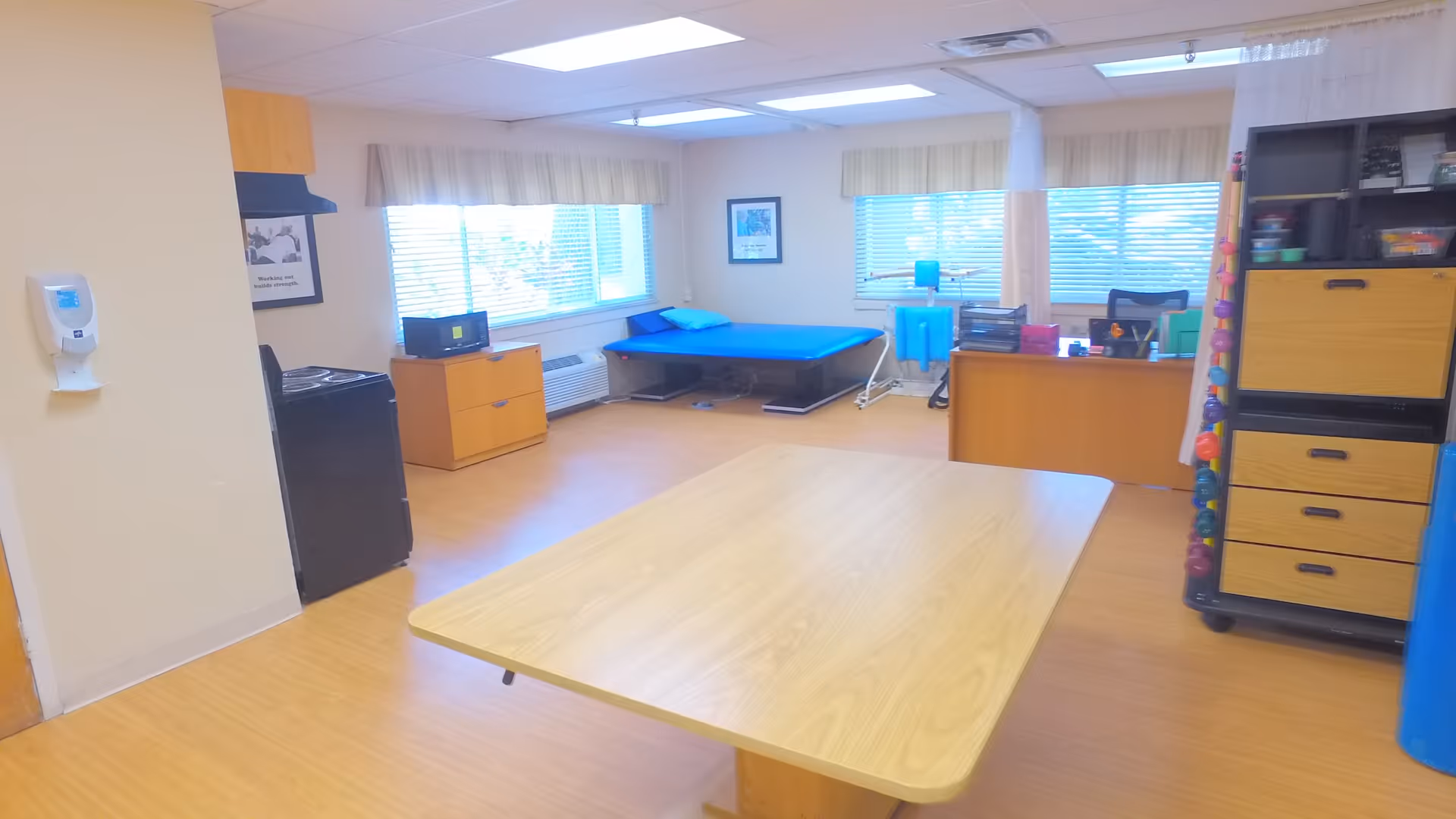 A bright room with wooden flooring featuring a large wooden table in the center, a blue cushioned examination or therapy table near the windows, a black stove, wooden cabinets, and a desk with office supplies. The room has large windows with blinds and light-colored walls.