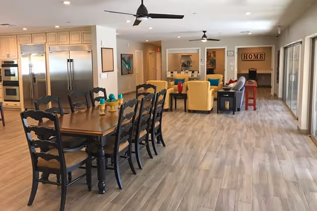 Open communal dining and living area with a long wooden dining table, yellow lounge chairs, and stainless-steel kitchen appliances.