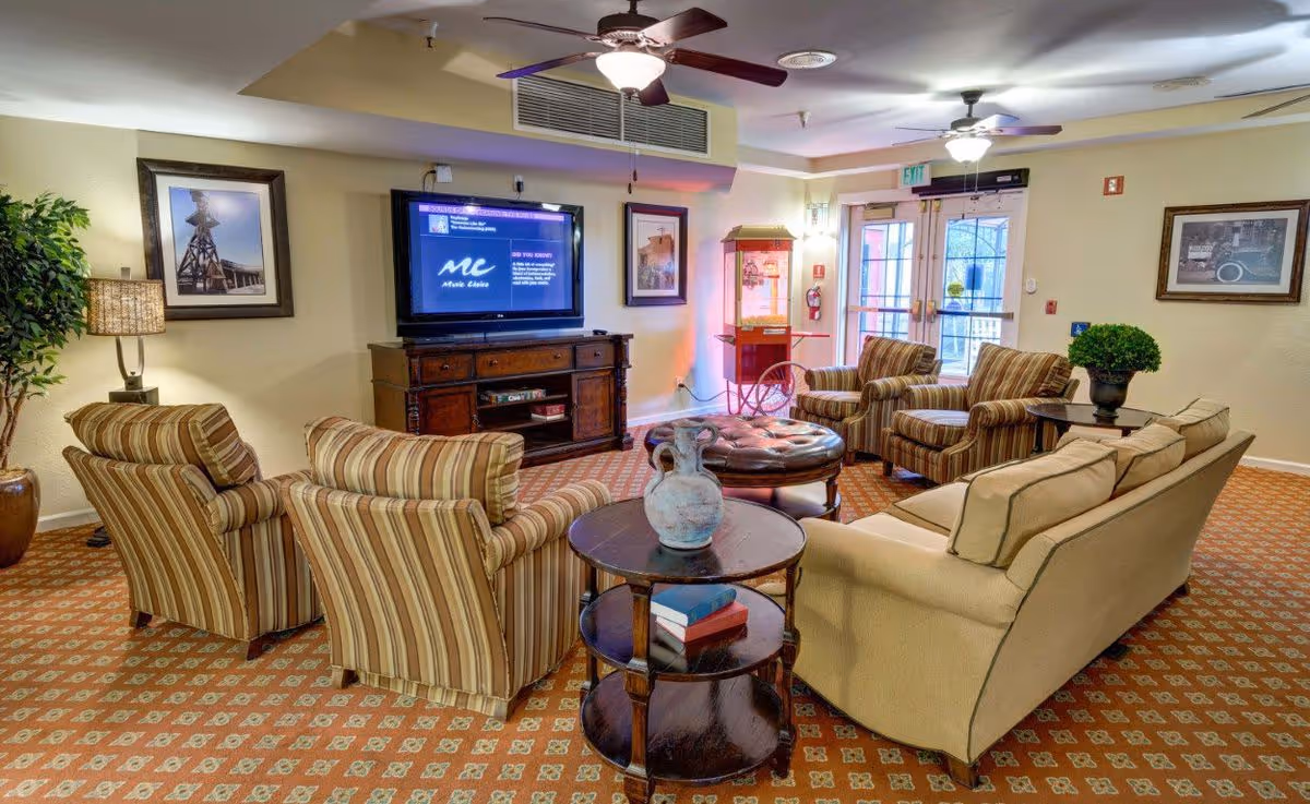 A comfortable senior living community lounge with sofas and striped armchairs arranged around a TV and coffee table.