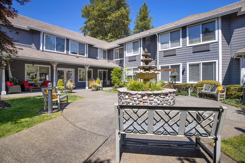 Outdoor courtyard area of a senior living facility with a multi-tiered stone fountain in the center, surrounded by benches and greenery. The building has gray siding with white trim and multiple windows, and there are trees in the background under a clear blue sky.