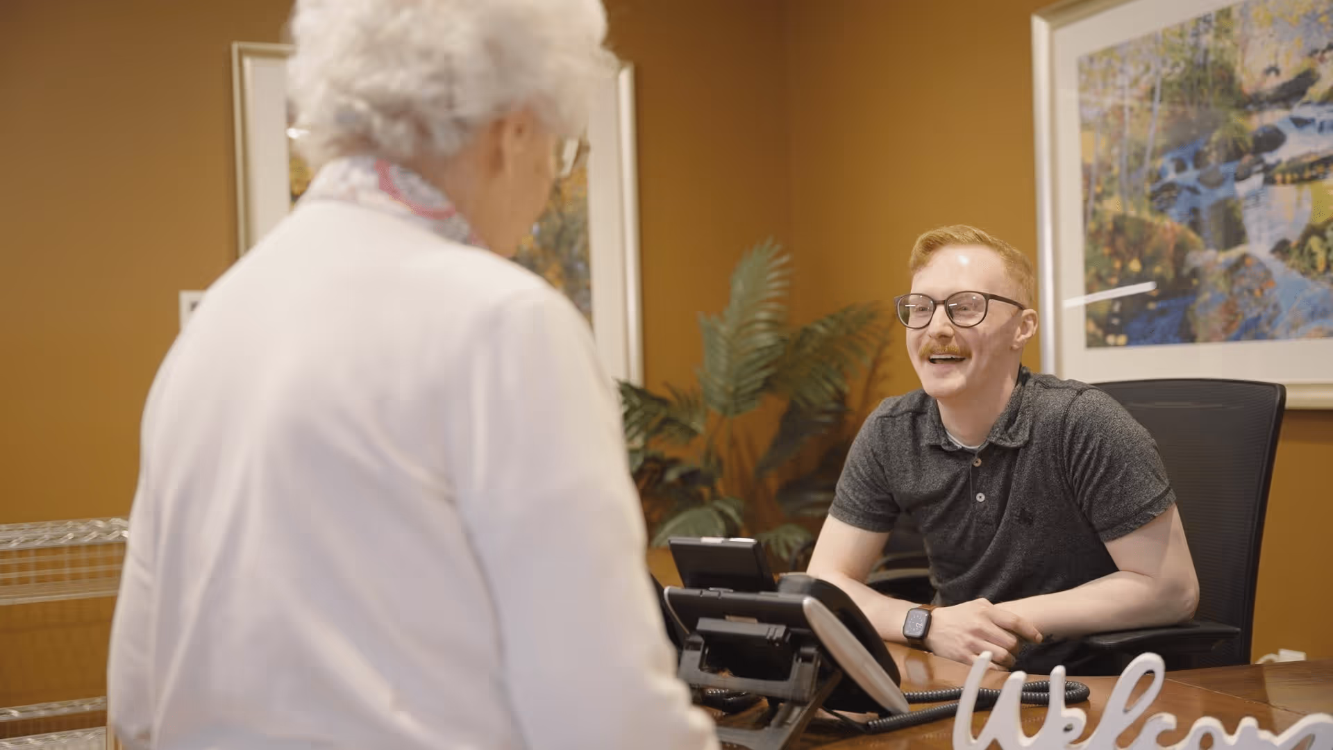 A young man with glasses and a mustache sits behind a desk smiling and talking to an elderly woman with white hair wearing a white jacket and a patterned scarf. The desk has a phone and a decorative 'Welcome' sign. The background features brown walls, framed artwork, and a green plant.