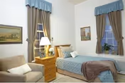 A cozy bedroom with a single bed covered in a blue bedspread and a brown throw blanket. There is a wooden nightstand with a lamp and a small clock next to the bed. Two windows with blue valances and beige curtains are visible, along with framed artwork on the walls. A beige armchair with a white pillow is positioned near the nightstand.