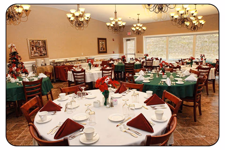A dining room in a senior living community with multiple round tables covered with white and green tablecloths. Each table is set with cups, plates, silverware, and folded napkins in red and white. There are red poinsettia centerpieces on the tables. The room has large windows, chandeliers, framed artwork on the walls, and a buffet setup along one side.