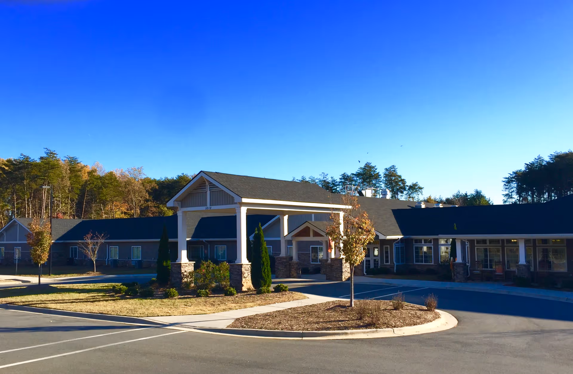Front entrance of a single-story senior living facility with a covered porte-cochere, landscaped islands, and a clear blue sky.