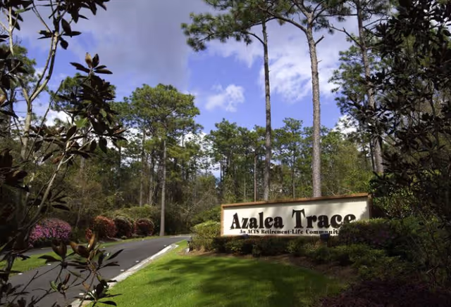 Entrance sign for Azalea Trace, an ACTS Retirement-Life Community, surrounded by greenery and tall pine trees under a partly cloudy sky.