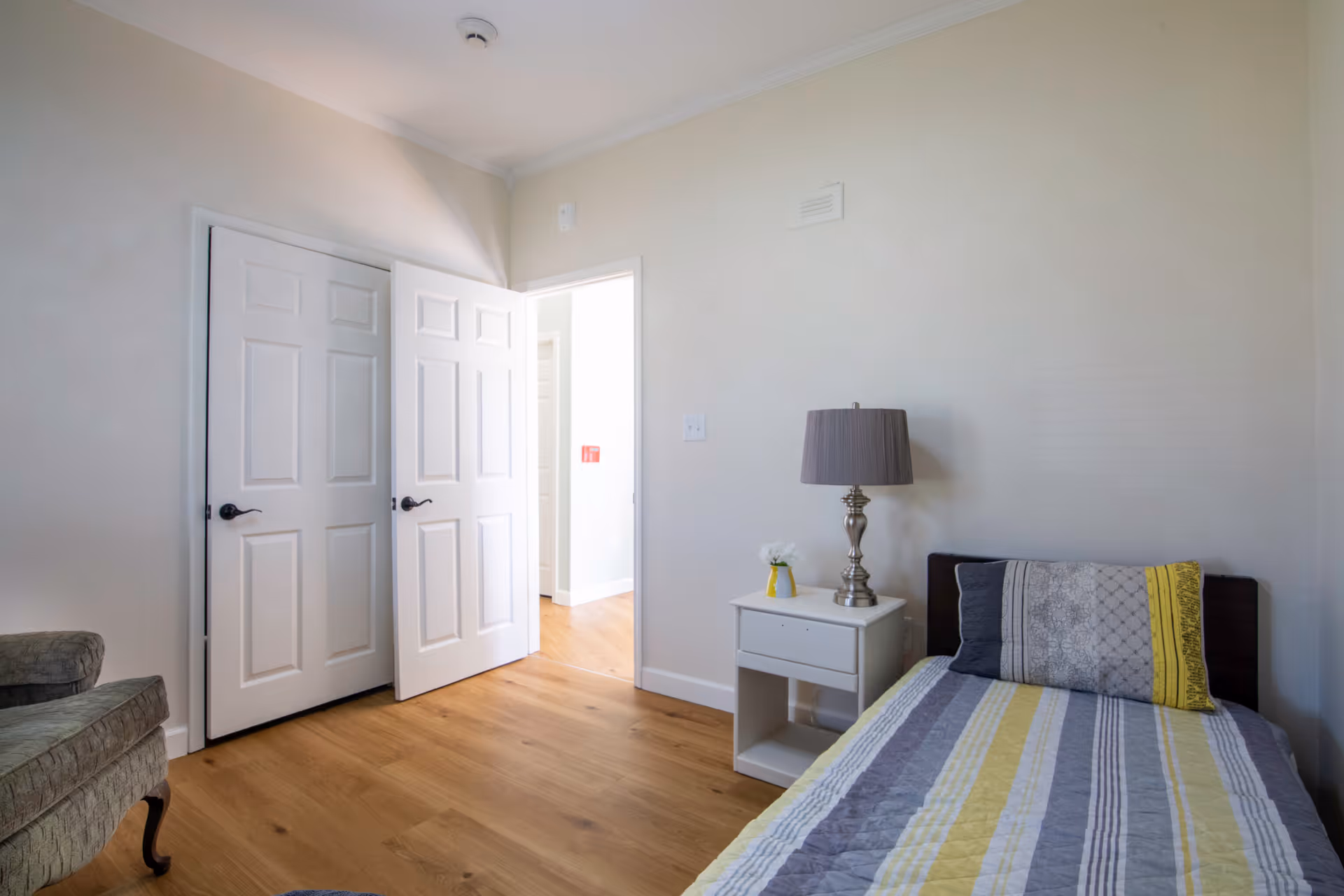 A simple bedroom with a single bed covered in a striped bedspread in shades of gray, yellow, and white. Next to the bed is a white nightstand with a silver lamp and a small vase with white flowers. The room has light-colored walls and wooden flooring. There is a partially open white door leading to another room, and a gray upholstered chair is partially visible on the left side.