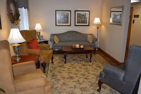 A cozy living room area with a patterned sofa, two armchairs, a coffee table with decorative items, two table lamps, a floor lamp, framed artwork on the wall, and a floral area rug on the floor.